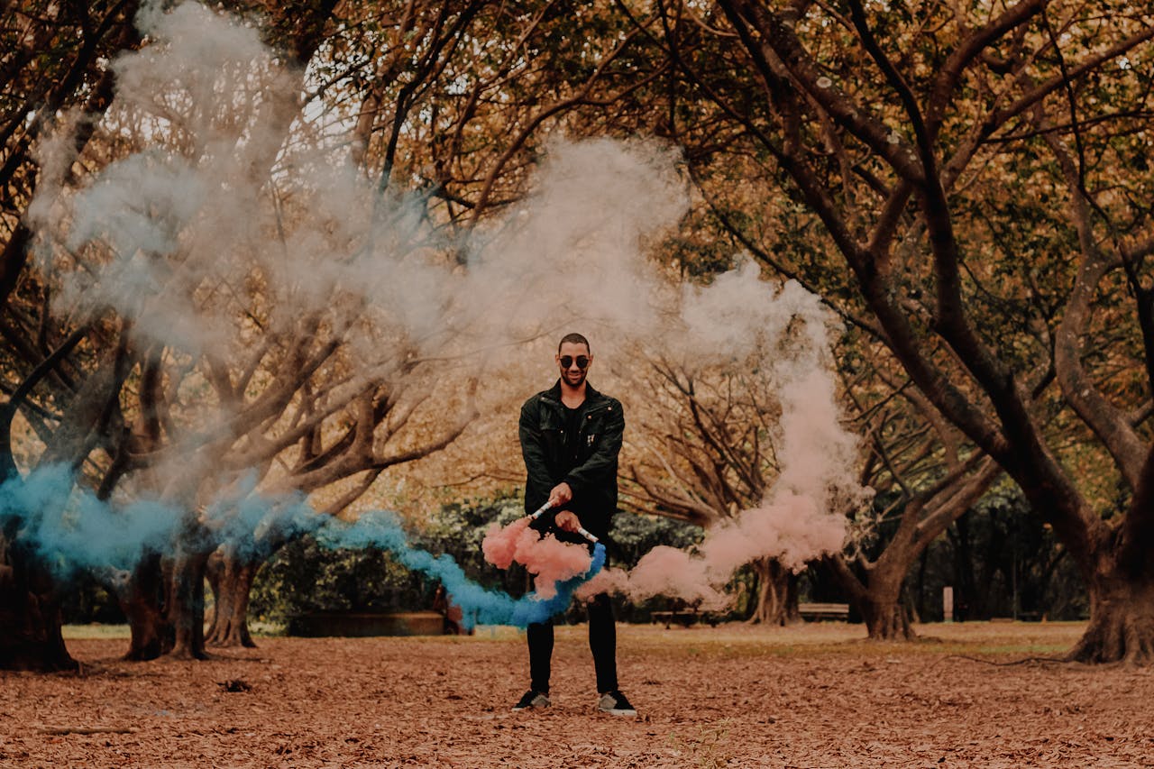 A man with sunglasses dispersing colorful smoke among autumn trees for a creative visual effect.
