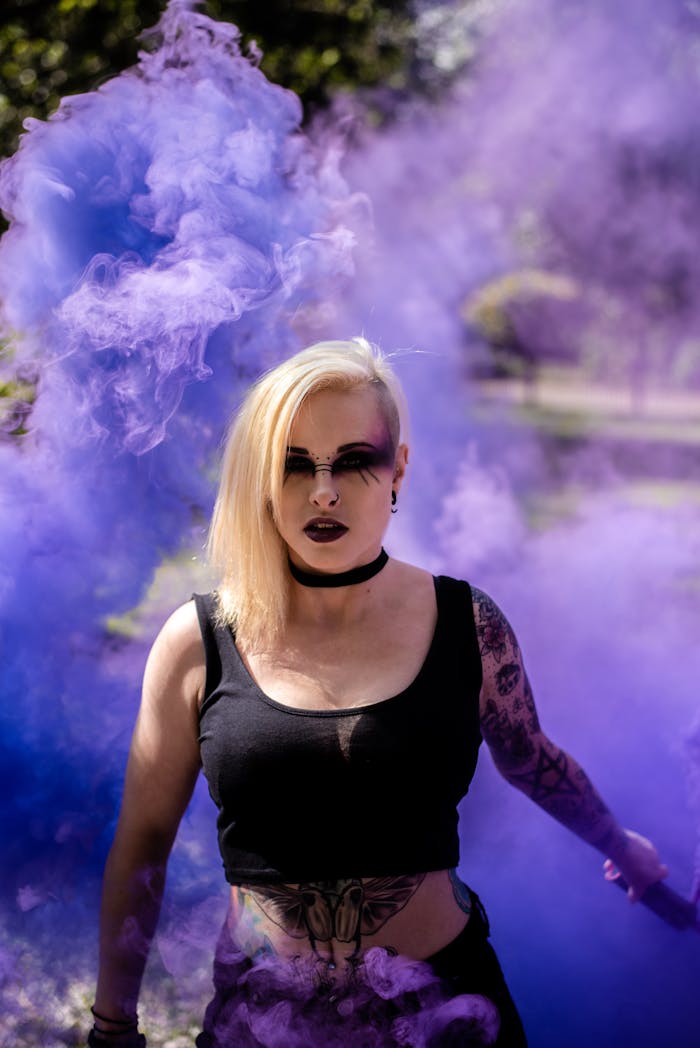 A blonde woman poses in a park surrounded by dramatic purple smoke under sunlight.